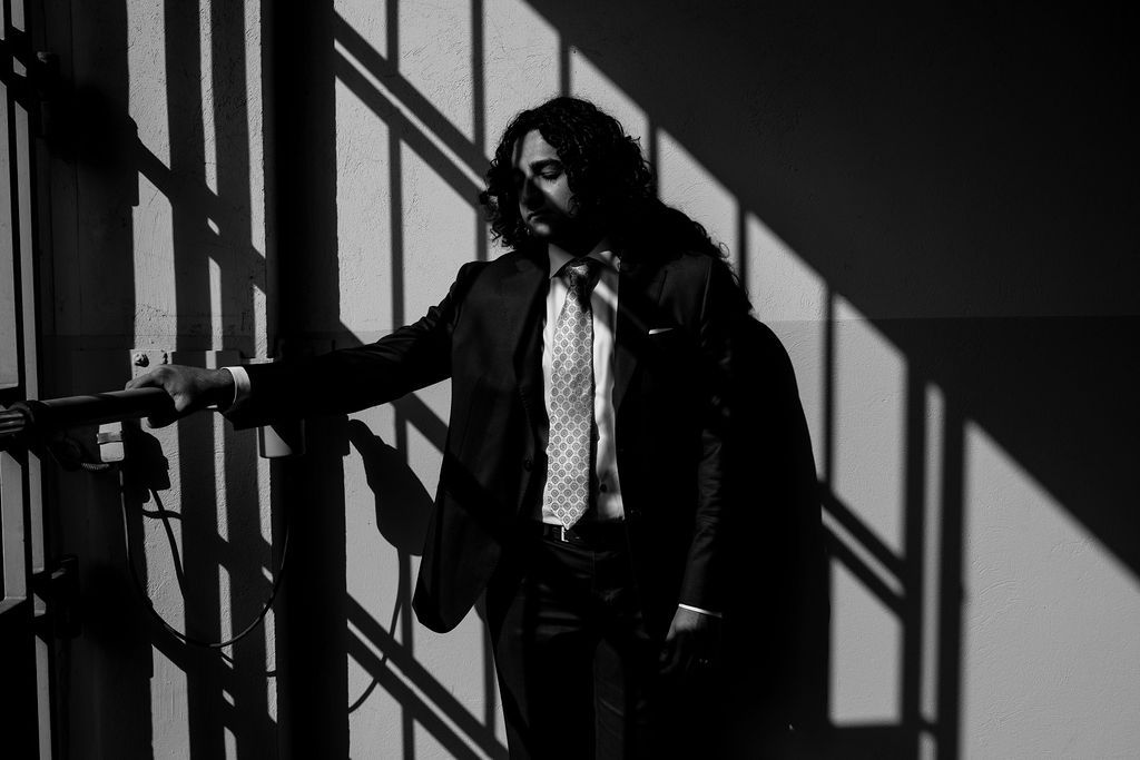 Black and white photo of a man in a suit and tie leaning against a wall with dramatic shadows.