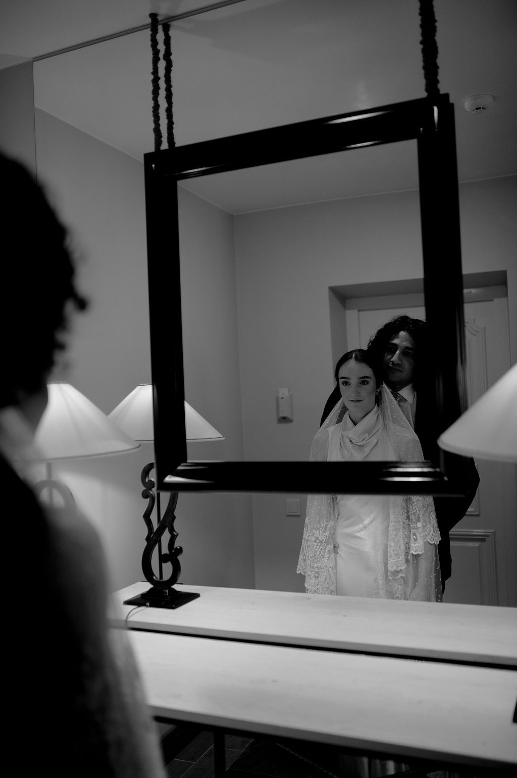 Black and white photo of a bride in white dress and veil reflected in an ornate hanging mirror with groom behind her.