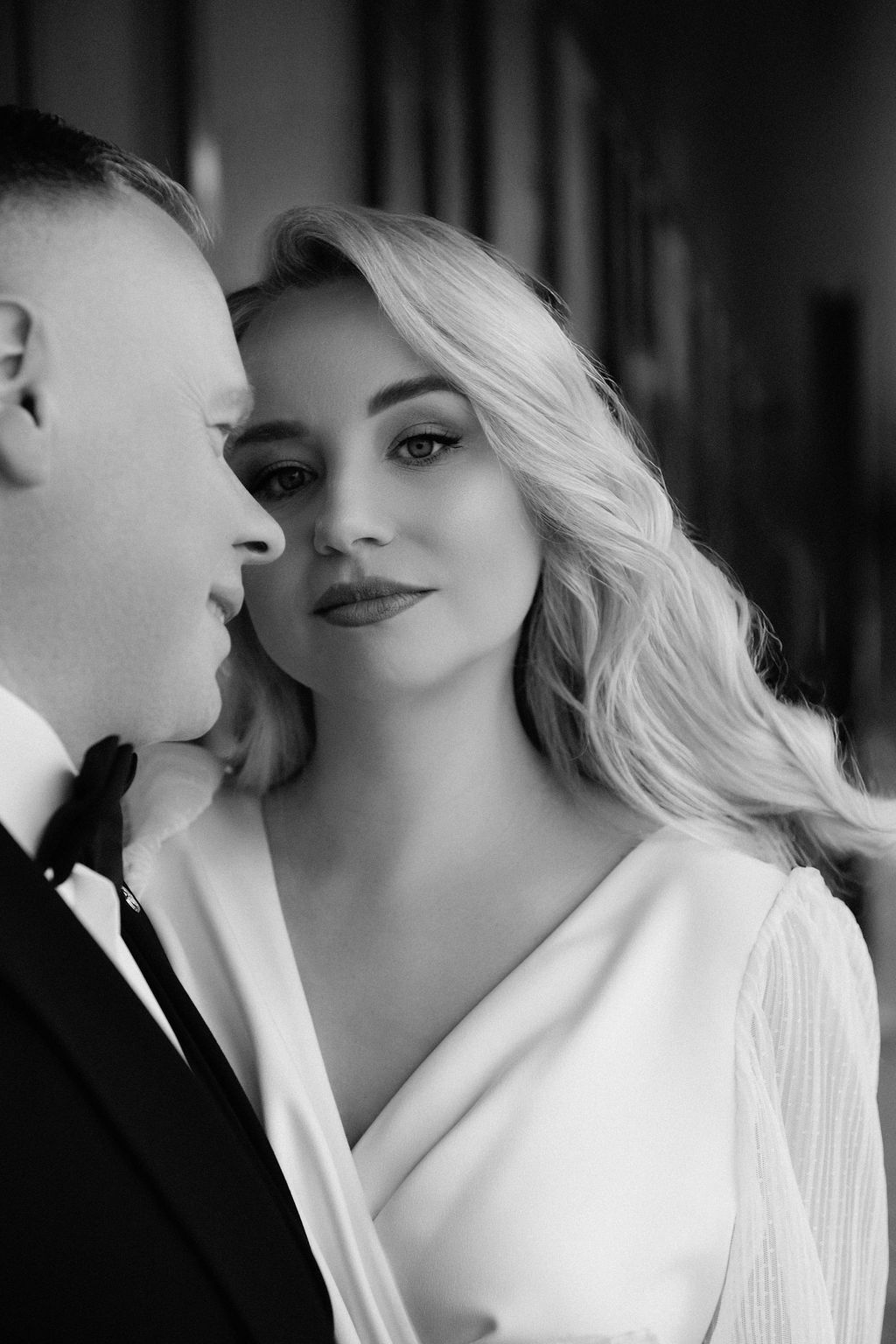 Black and white wedding portrait of elegant blonde bride in white gown gazing at camera beside groom in tuxedo.