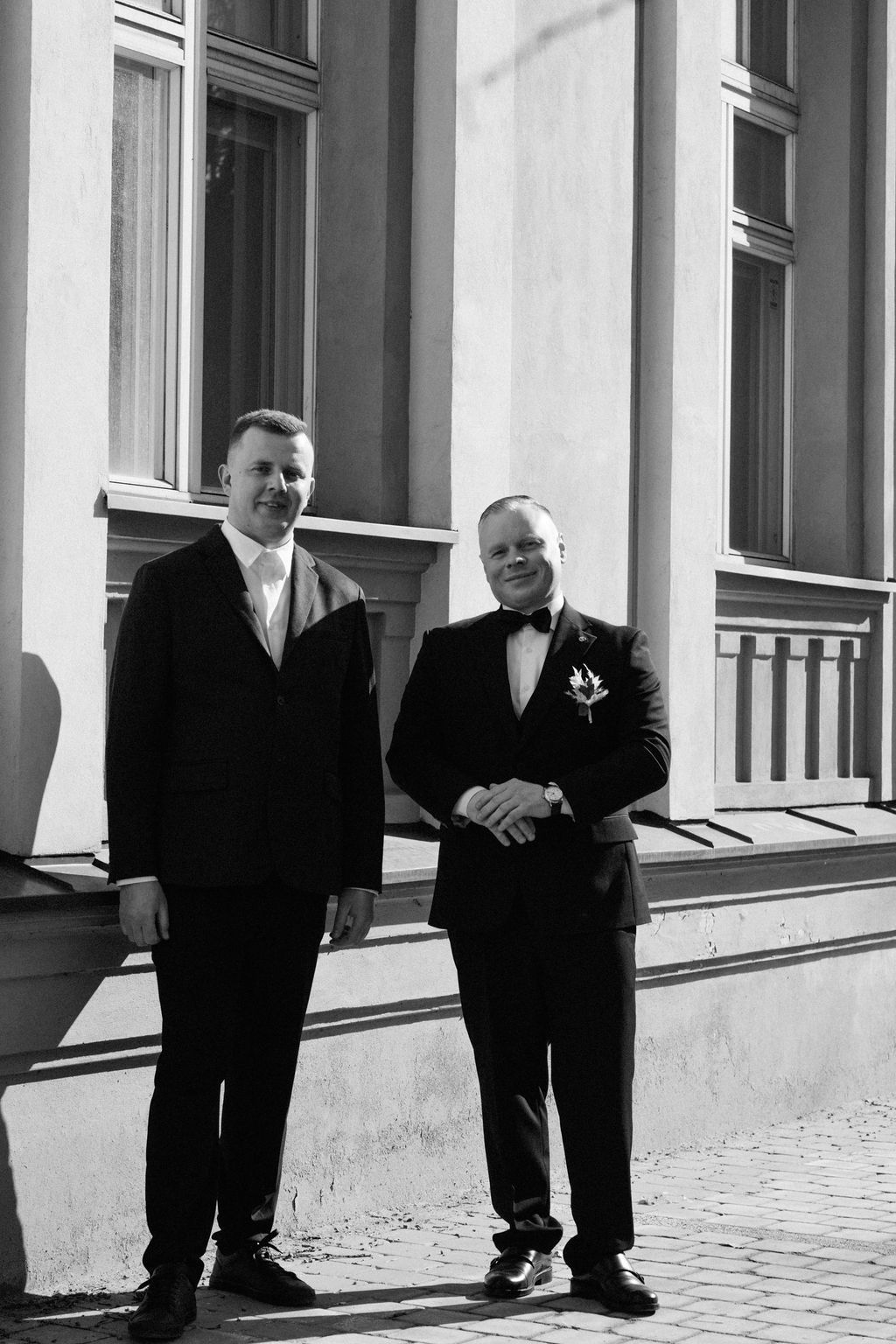 Two men in formal suits posing outside a classic building in black and white wedding photo.
