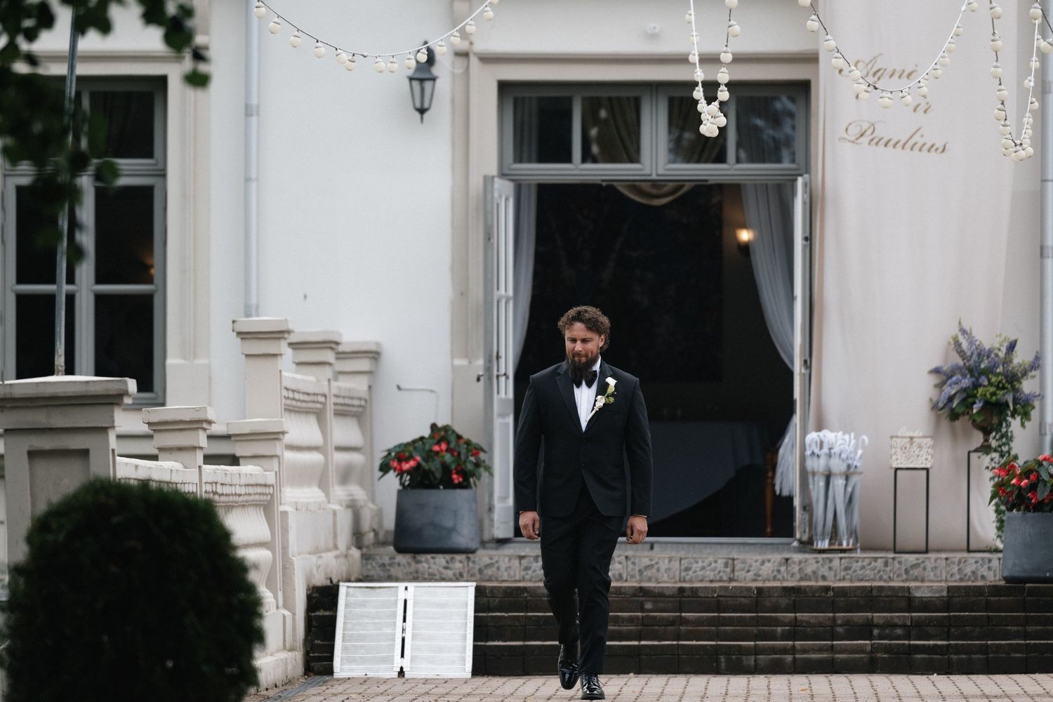 Groom in black tuxedo standing outside elegant white wedding venue with string lights and decorative entrance.