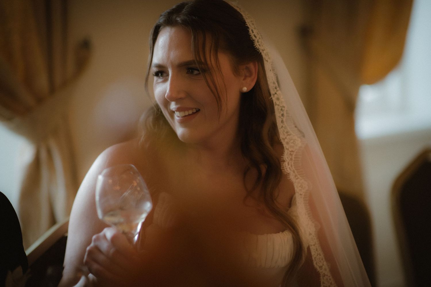 Smiling bride wearing lace veil and strapless gown holding champagne glass indoors.