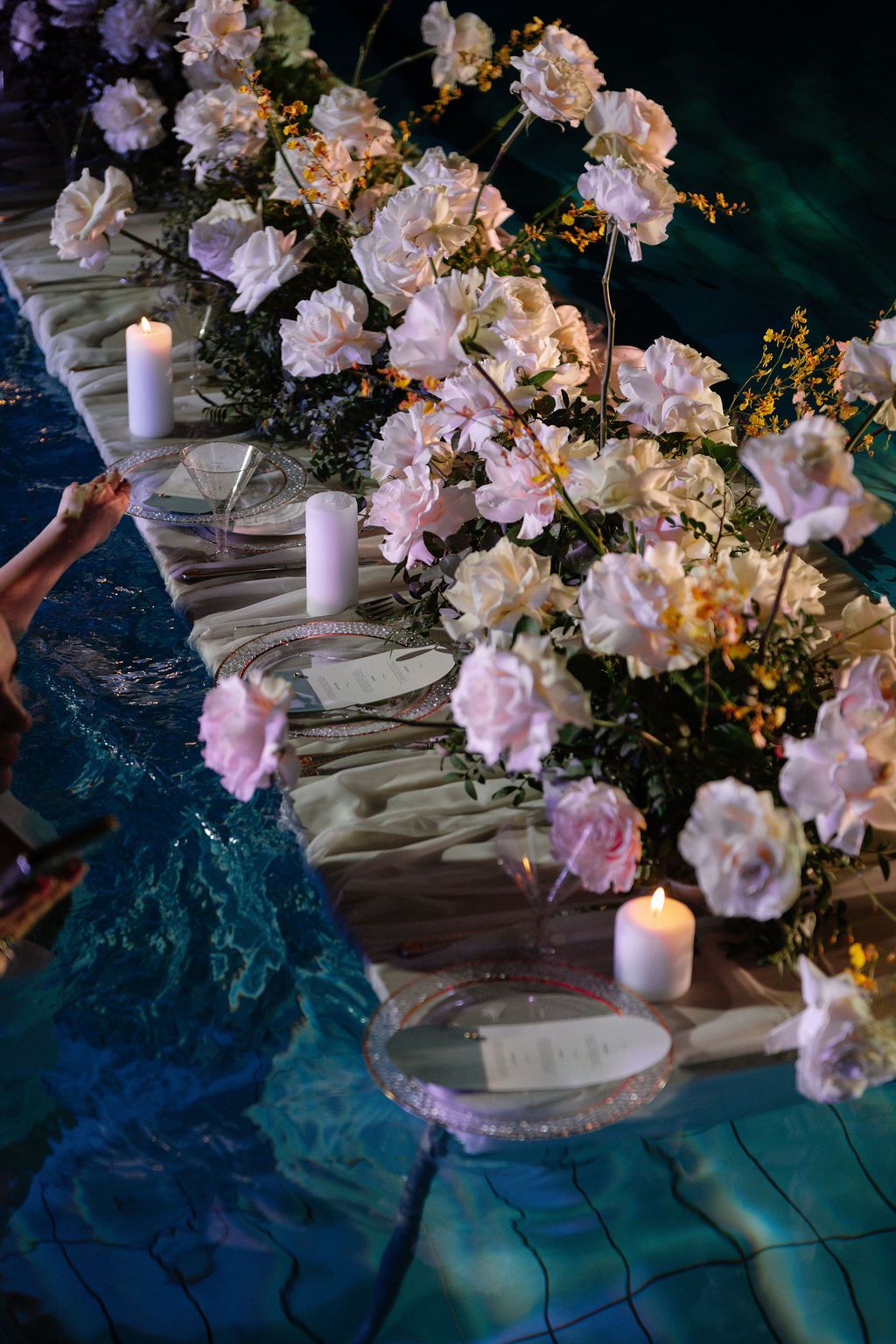 Pale pink and white roses with lit candles and place settings arranged on a floating table over a glowing blue pool at night.