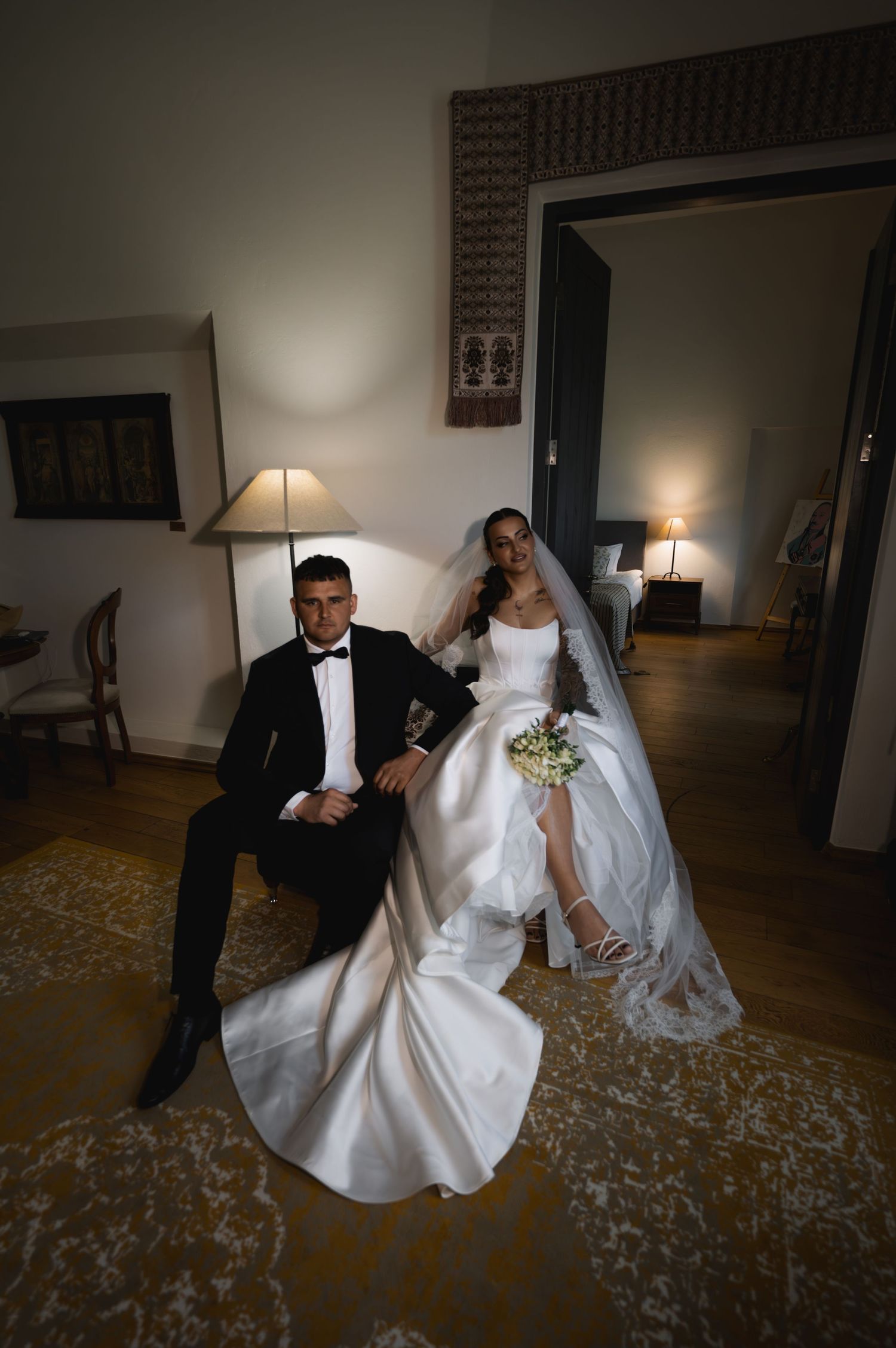 Bride in a flowing white gown and veil sits beside a groom in a black tuxedo and bow tie in an elegant, softly lit room.