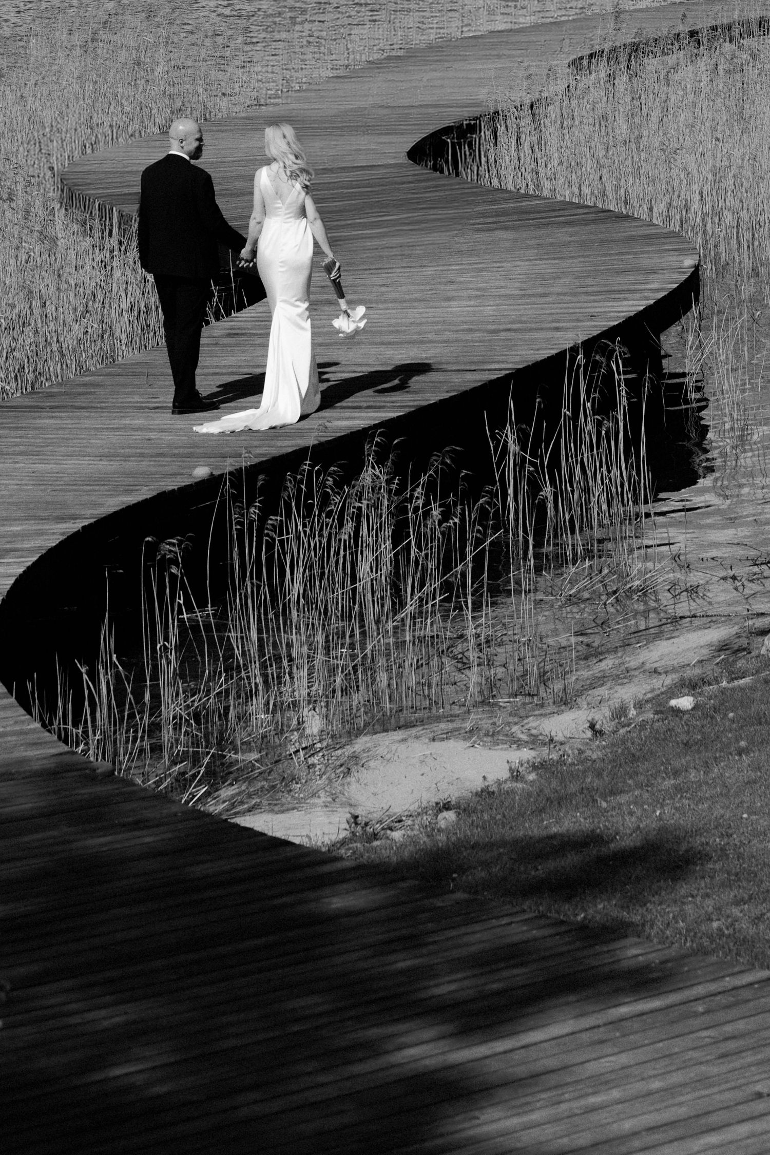 Bride in a white gown and groom in a dark suit walk hand in hand along a curving wooden boardwalk through tall reeds in black and white.