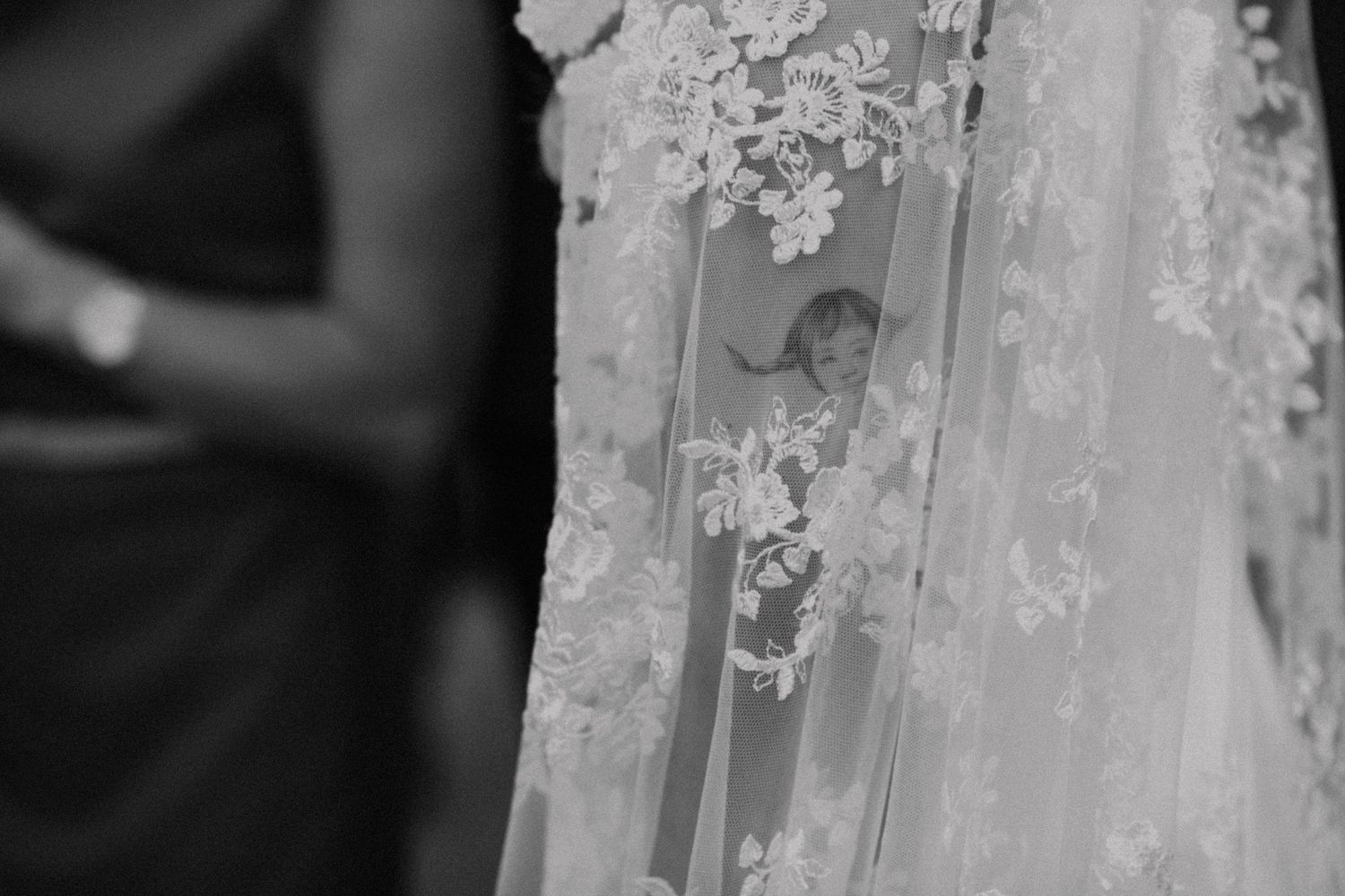 Black and white close-up of a delicate lace wedding veil with floral embroidery, with a blurred guest in the background.