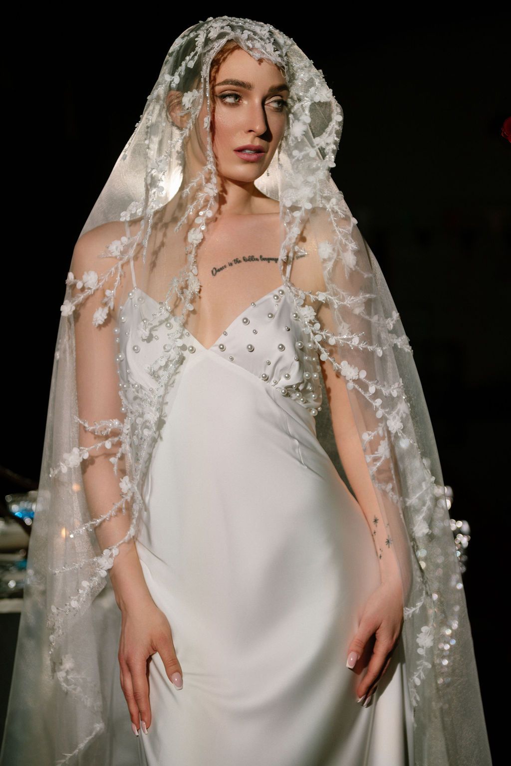 Bride in a white satin slip wedding dress with a floral-embellished sheer lace veil draped over her head and shoulders.