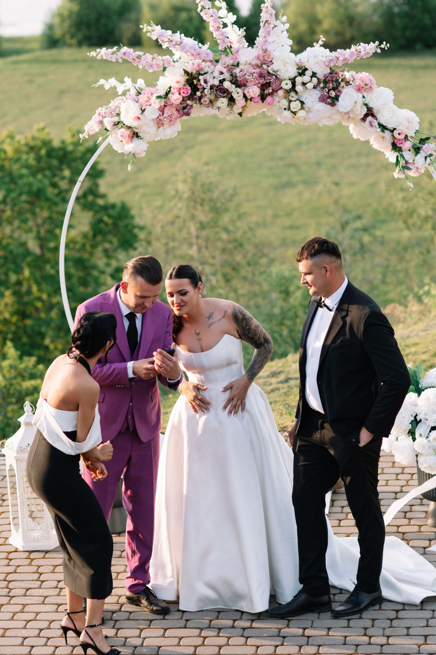 Wedding ceremony under a floral arch with a bride in a white gown, an officiant in a purple suit, a groomsman and a woman in black.