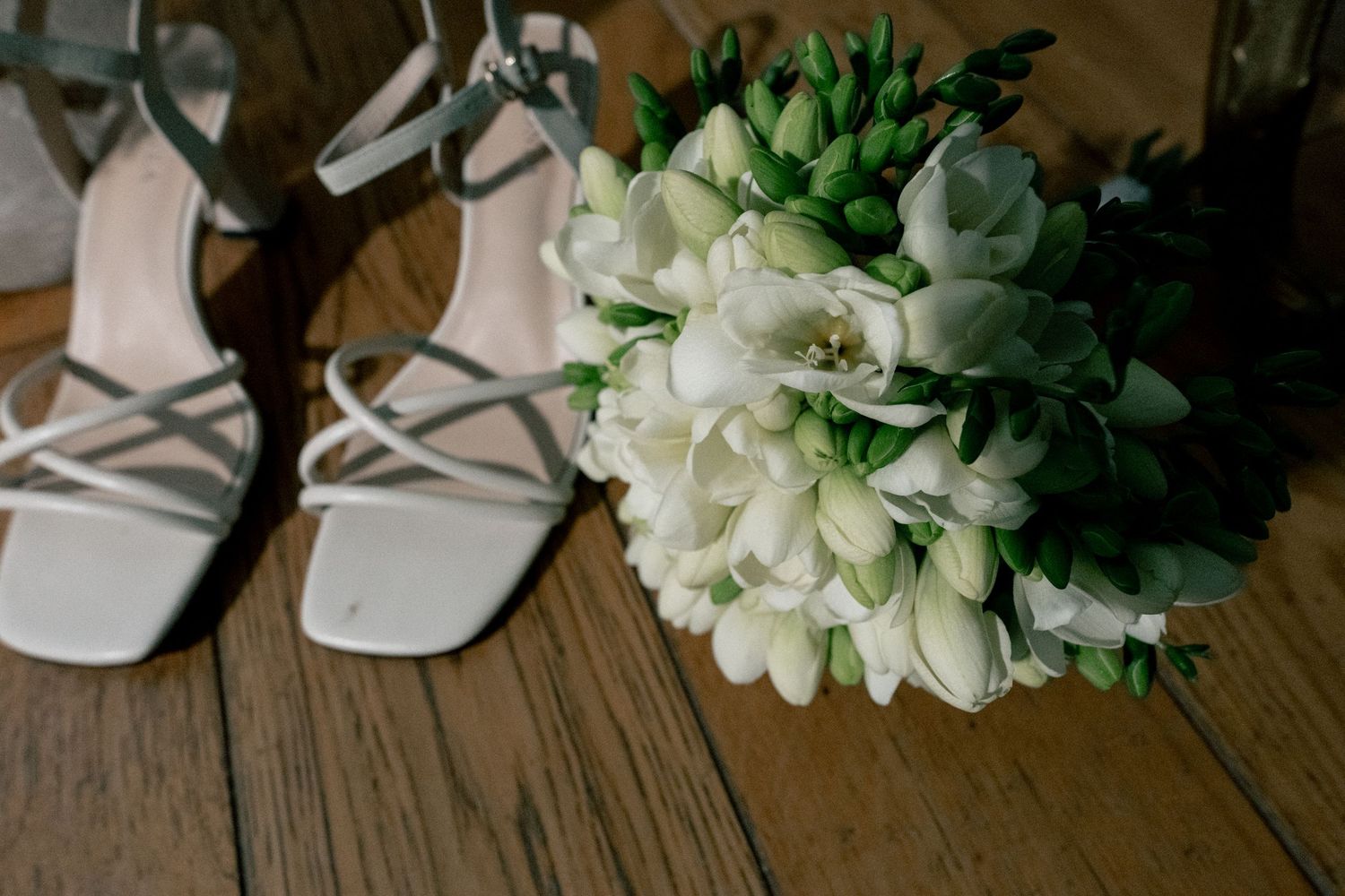 White strappy heeled sandals beside a bridal bouquet of white freesias and green buds on a wooden floor.
