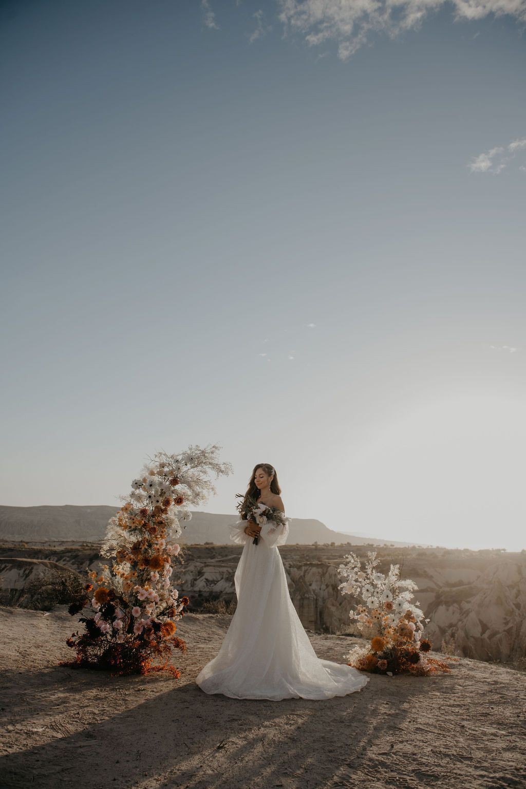 Bride in flowing white gown standing beside floral arch with autumn blooms at scenic cliffside wedding venue.