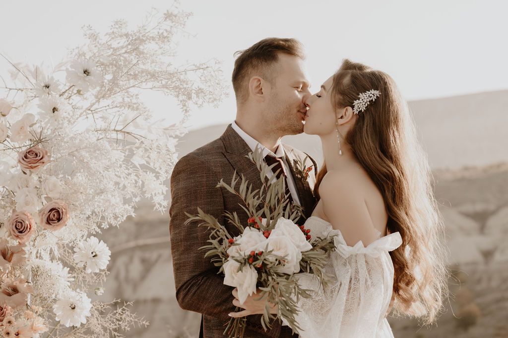 Bride and groom share a romantic kiss surrounded by white and blush floral arrangements on their wedding day.