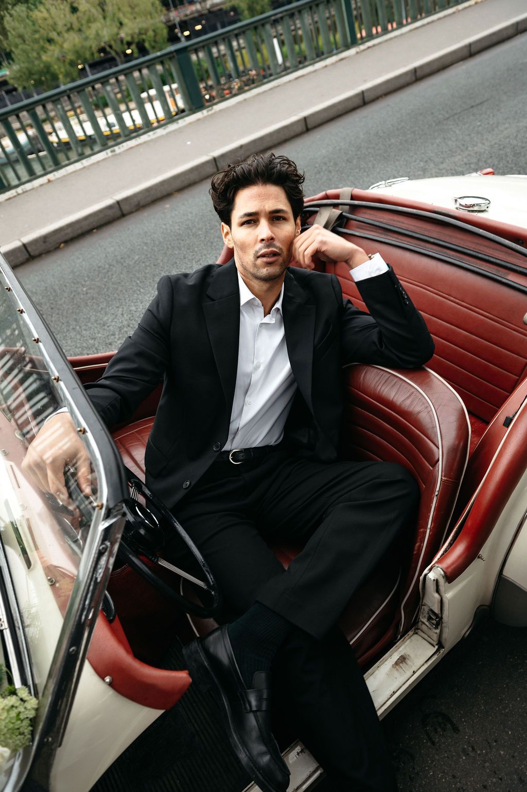 A stylish man in a black suit sits confidently in a vintage convertible car with red leather interior.