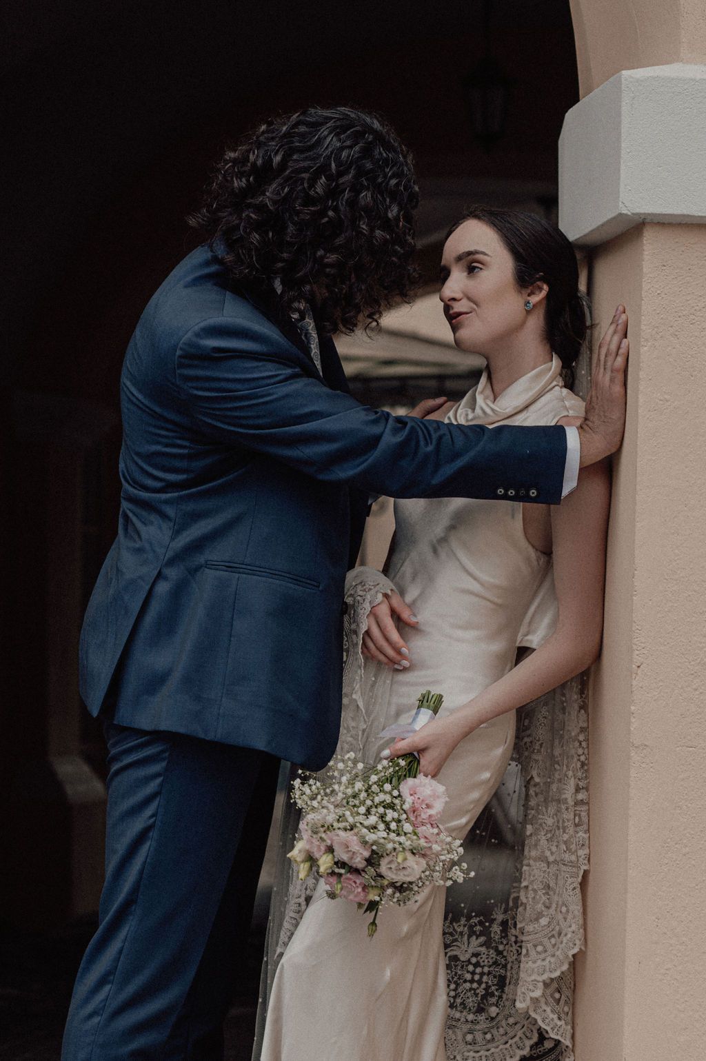 A groom in a navy suit leans toward his bride in a vintage lace gown holding a pink bouquet against a column.