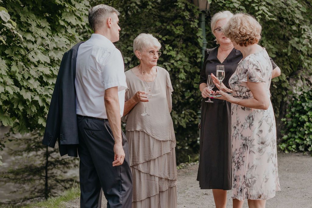 Elegantly dressed seniors socializing outdoors at a wedding reception, holding wine glasses near ivy-covered stone wall.