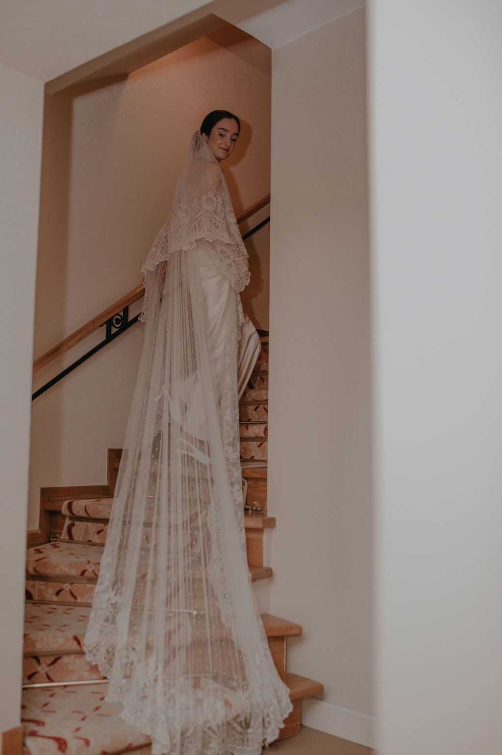 Bride in a stunning lace wedding veil and gown standing on an elegant staircase.