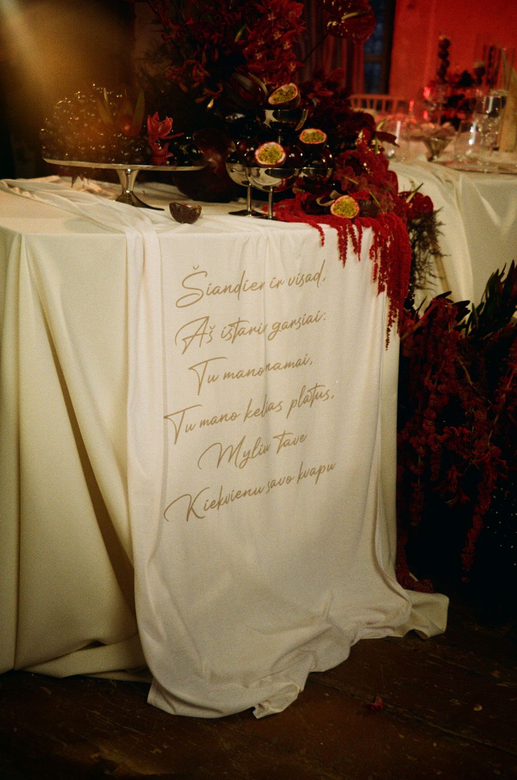 Elegant wedding table with white cloth banner featuring gold Lithuanian love poem, red floral decor.