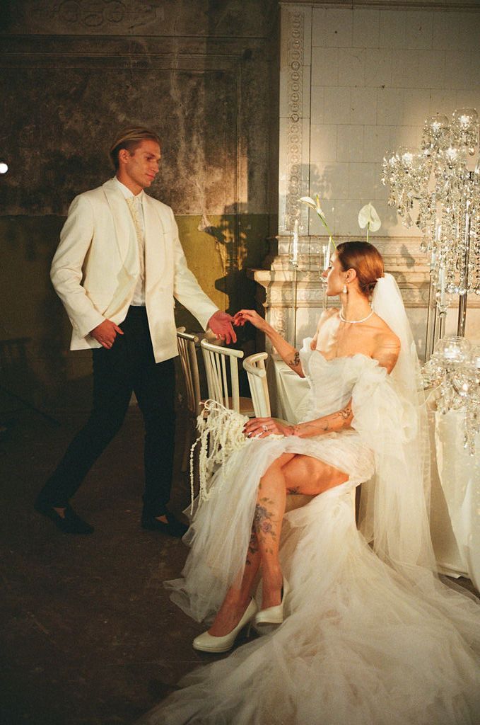 Elegant bride in white tulle gown seated while groom in ivory suit stands beside her at a glamorous wedding venue.