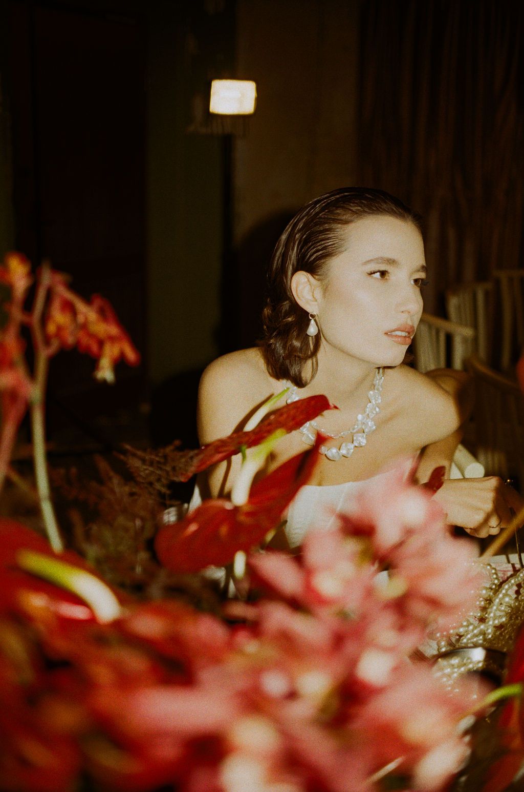 Woman in elegant jewelry surrounded by red tropical flowers under warm studio lighting.