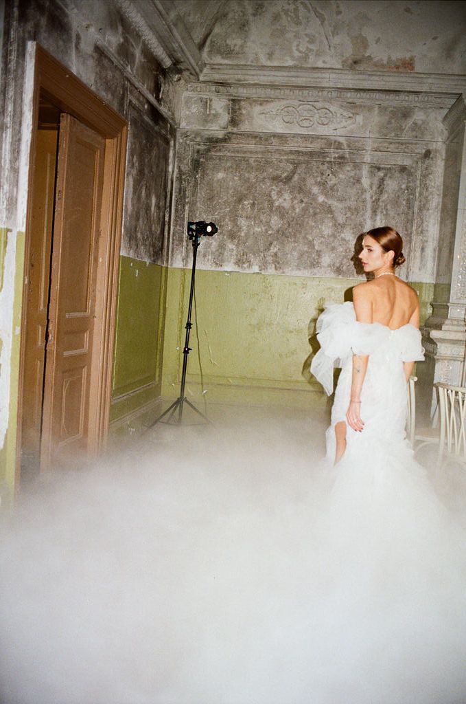Bride in off-shoulder white gown stands in misty ornate hall during dreamy bridal photoshoot.