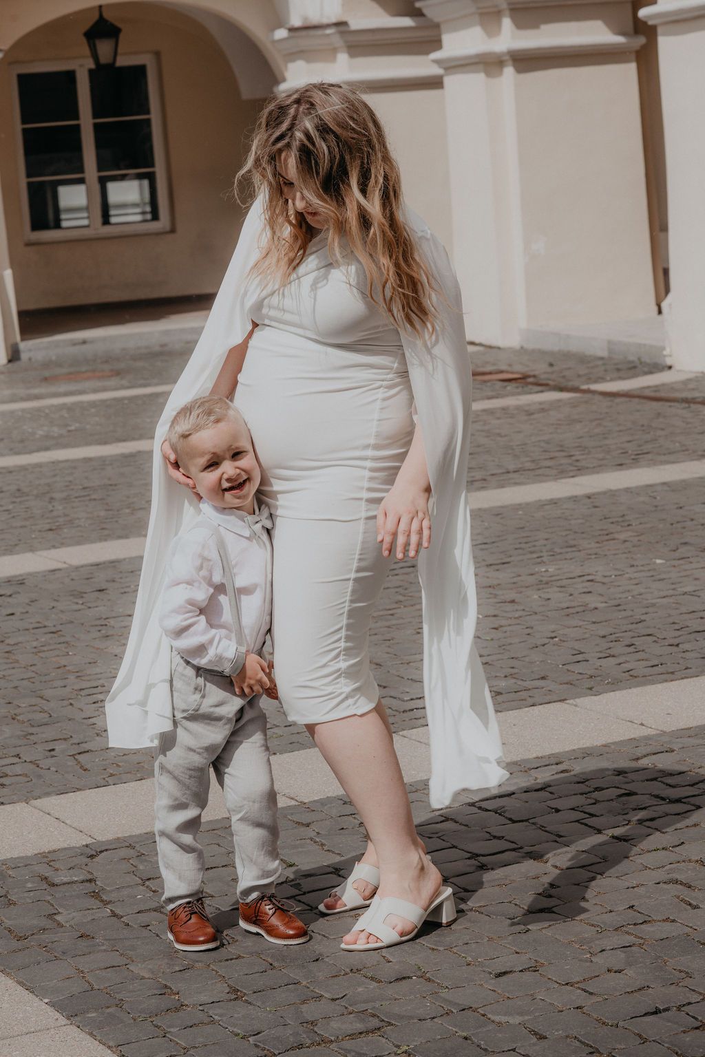 Pregnant woman in white dress with young toddler boy in suit smiling on cobblestone plaza.