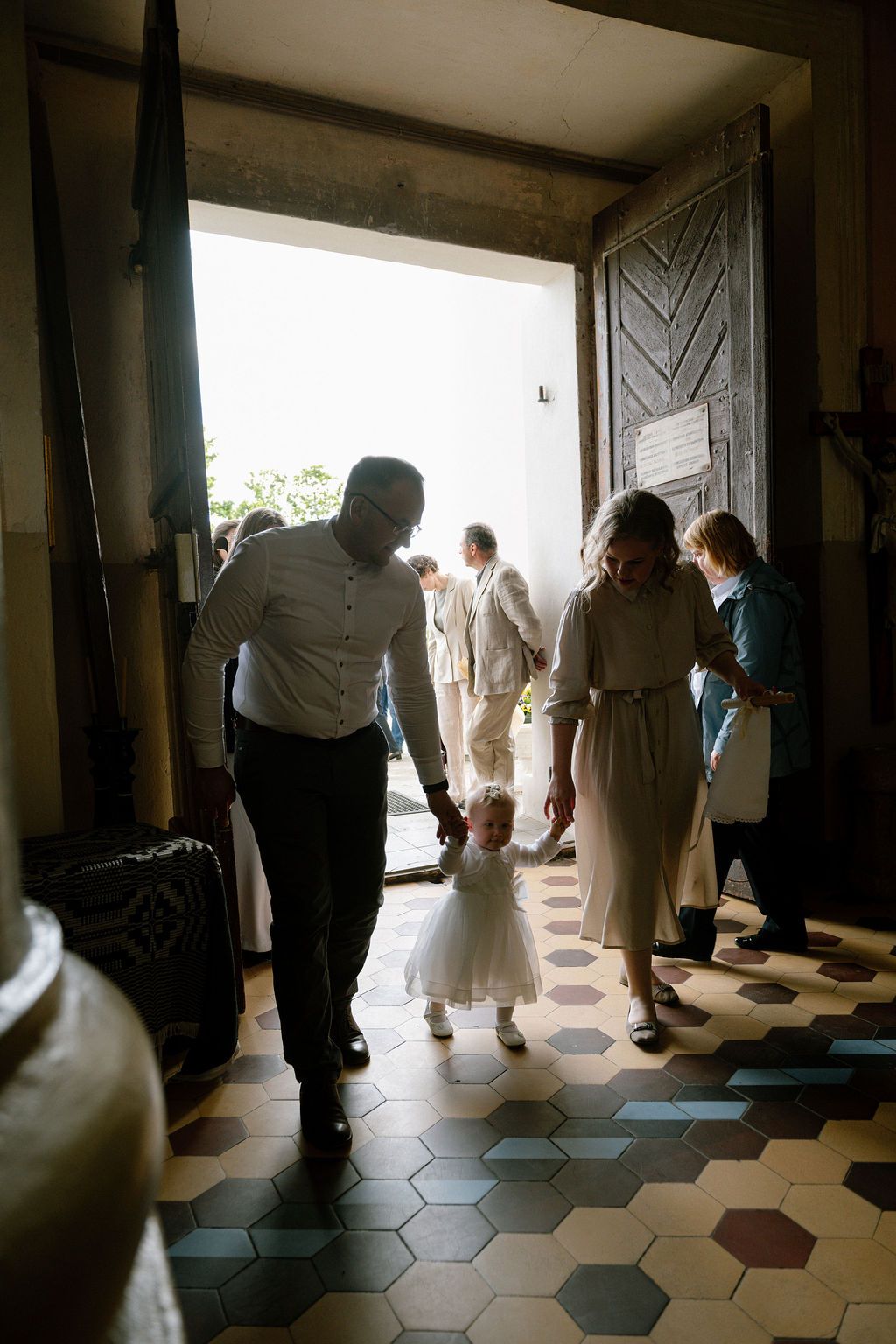 A couple guides a young flower girl in a white dress through a sunlit church doorway on a wedding day.