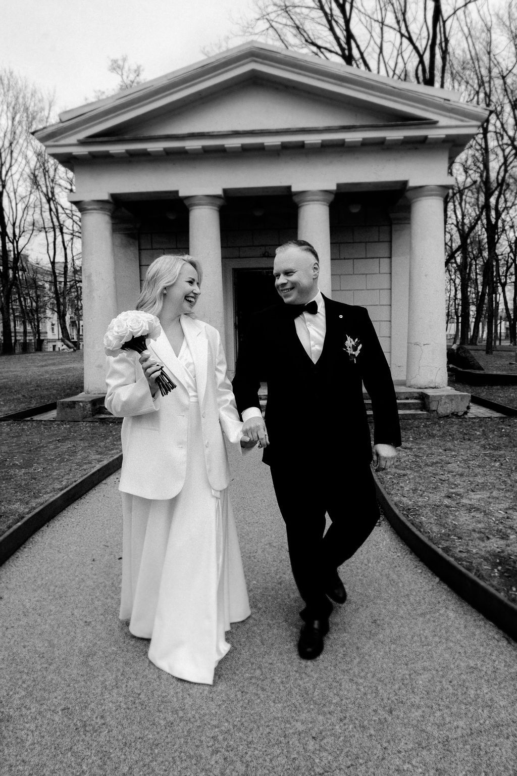 Black and white wedding photo of smiling bride and groom walking hand in hand before a classical columned building.