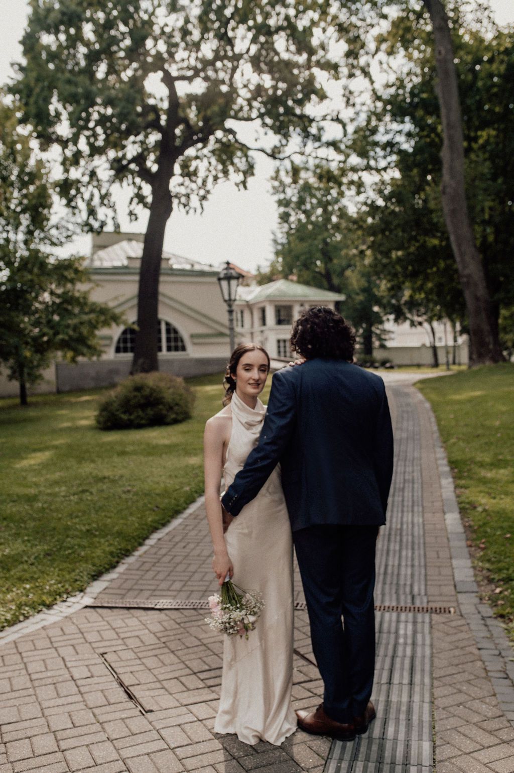 A bride in a white gown holds flowers while walking with a groom in a navy suit on a brick path in a park.