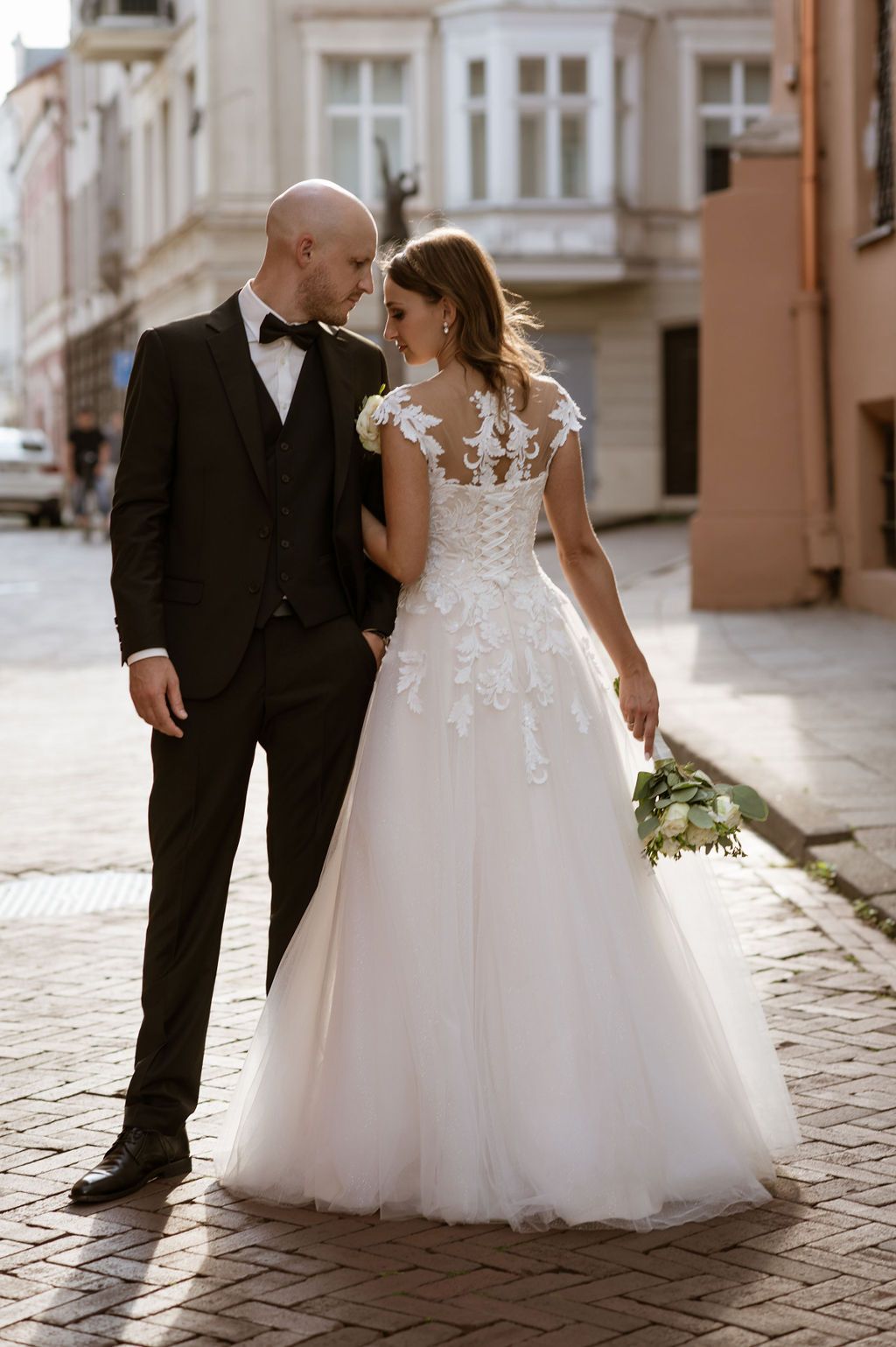 Bride and groom walking on cobblestone street, she in lace A-line gown with bouquet, he in black tuxedo.