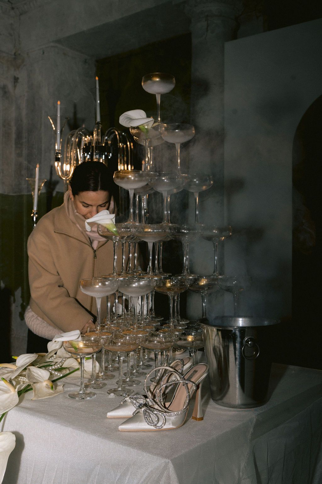 Woman arranging a champagne tower with coupe glasses at an elegant event, with heels and floral decor on a white tablecloth.