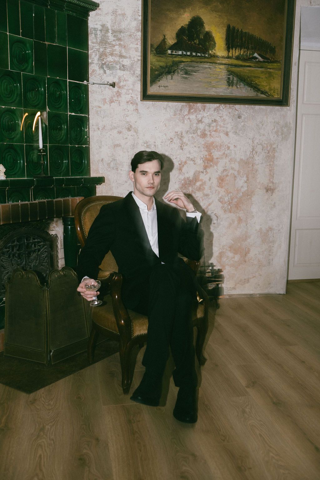 Sophisticated young man in black tuxedo seated in antique chair beside green tiled fireplace with landscape painting.