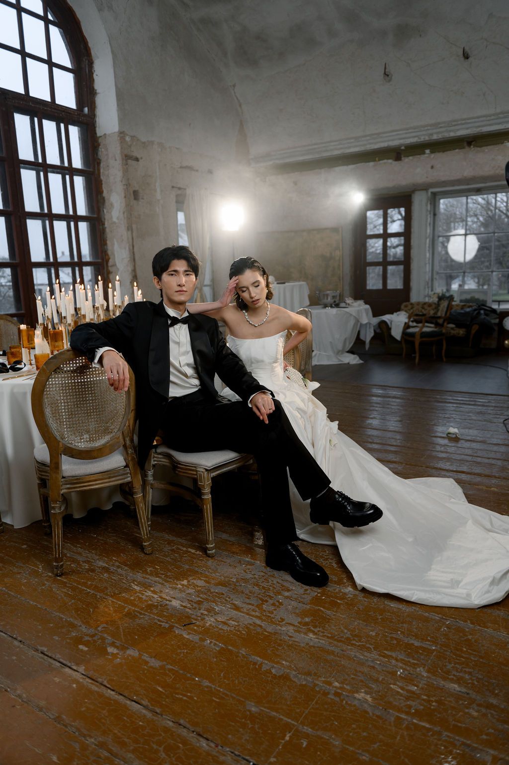 Wedding couple poses in rustic venue, groom in black tuxedo seated in ornate chair with bride in white gown leaning beside him.