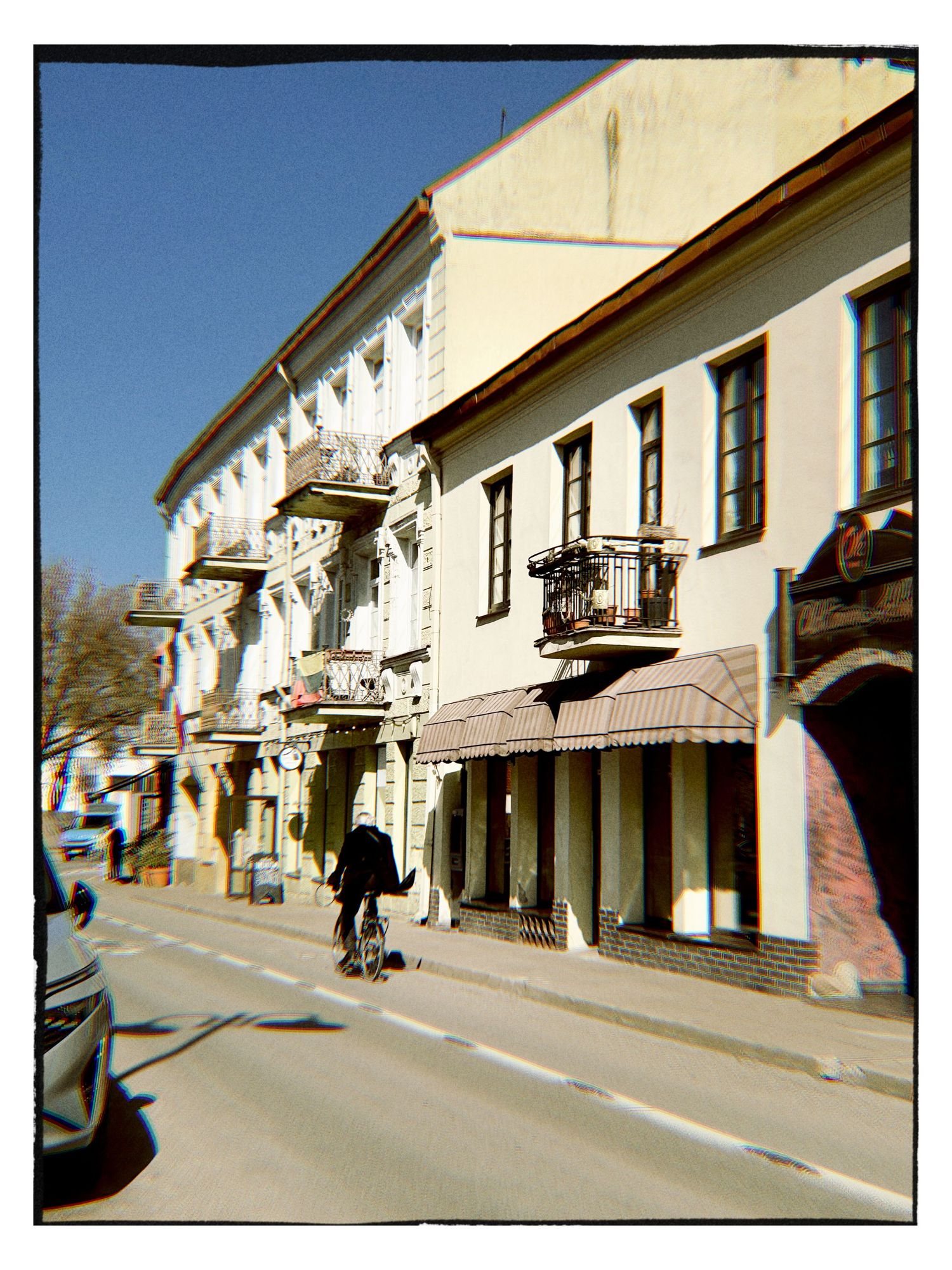 A cyclist rides past historic European buildings with balconies on a sunny small-town street.