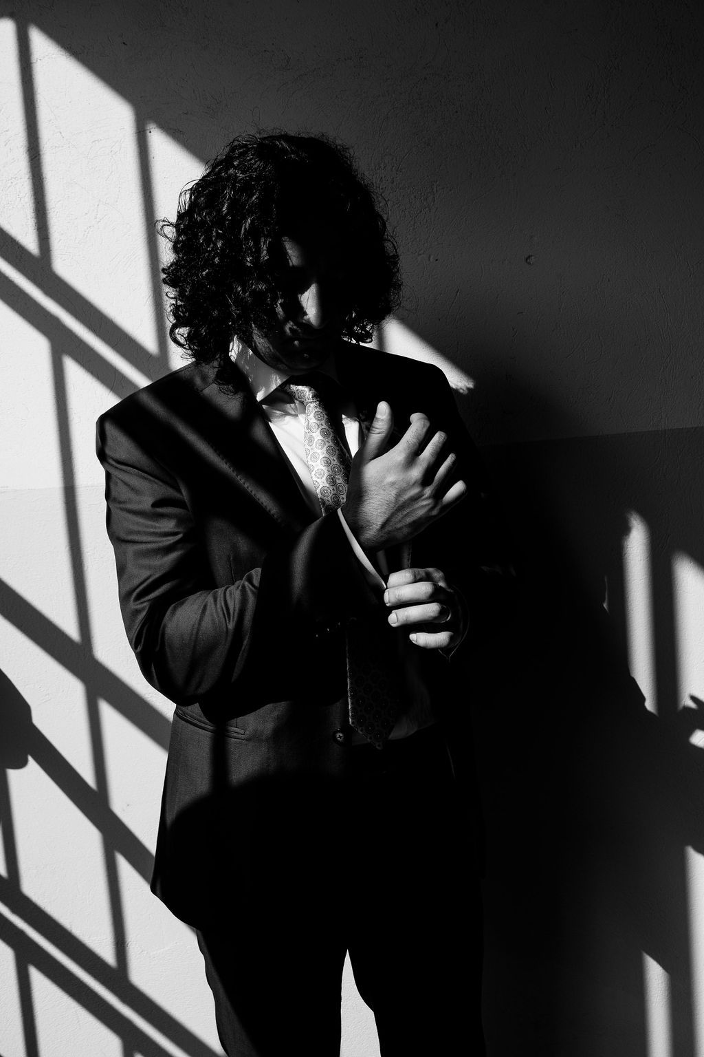 Black and white portrait of a curly-haired man in a suit adjusting his sleeve with dramatic window shadows.