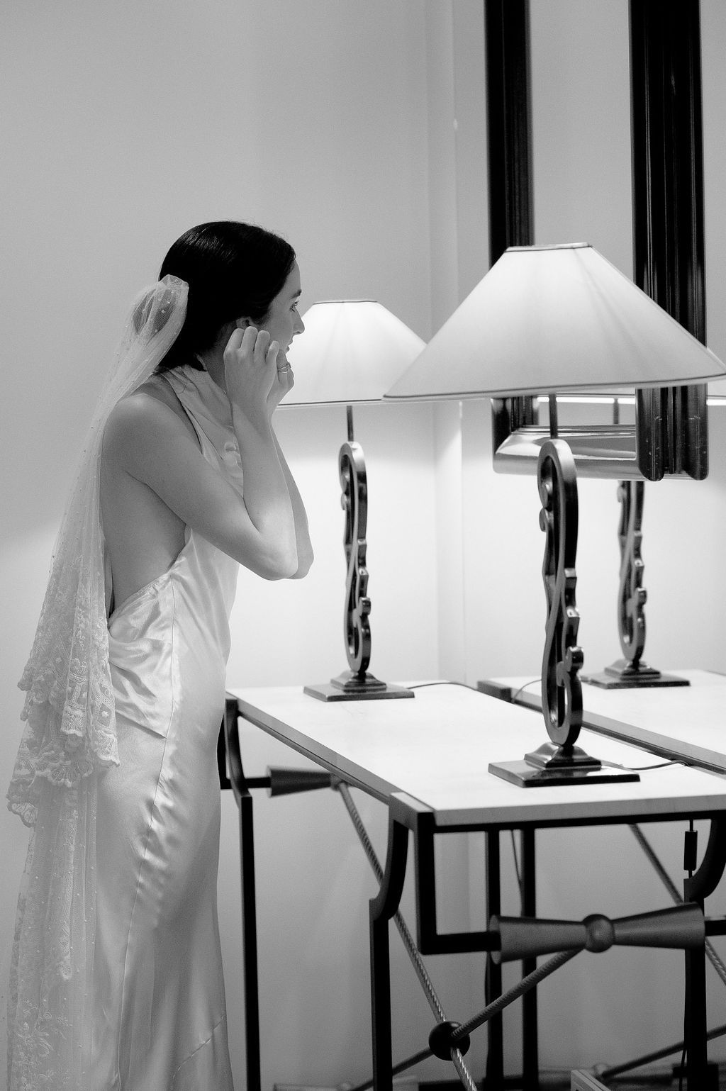 Black and white photo of a bride in a satin gown and veil standing by a table with two elegant lamps.