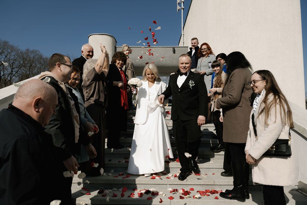 Newlywed couple walks through rose petal shower surrounded by cheering guests outside a wedding venue.