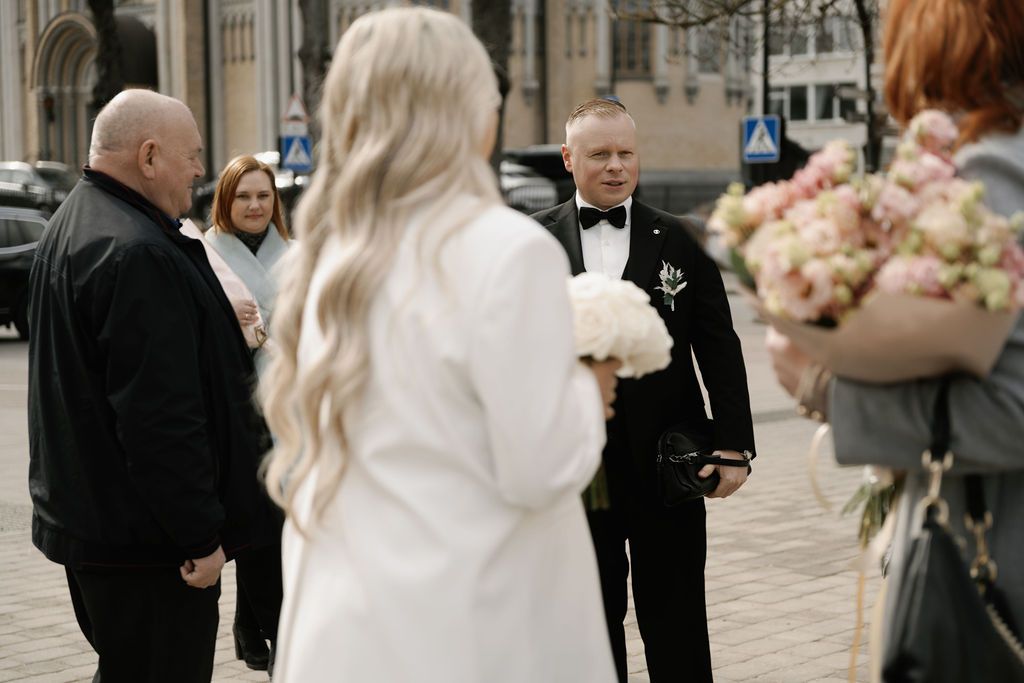 A bride in white holding a bouquet faces a groom in a tuxedo outdoors at a wedding ceremony.