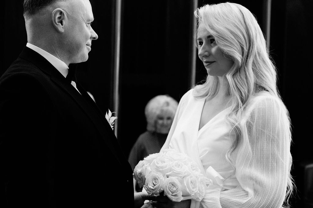 Black and white photo of bride holding white roses gazing at groom during wedding ceremony.