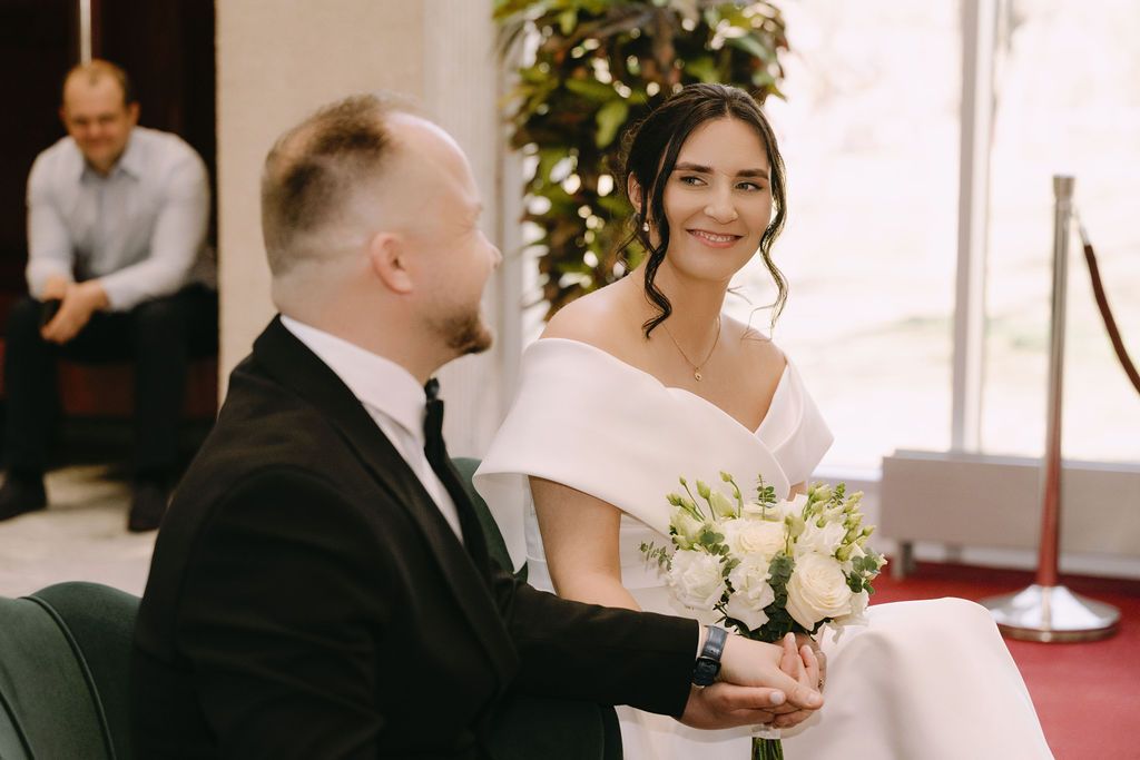 A smiling bride in a white off-shoulder gown holds flowers while a groom in a black suit holds her hands at a wedding ceremony.