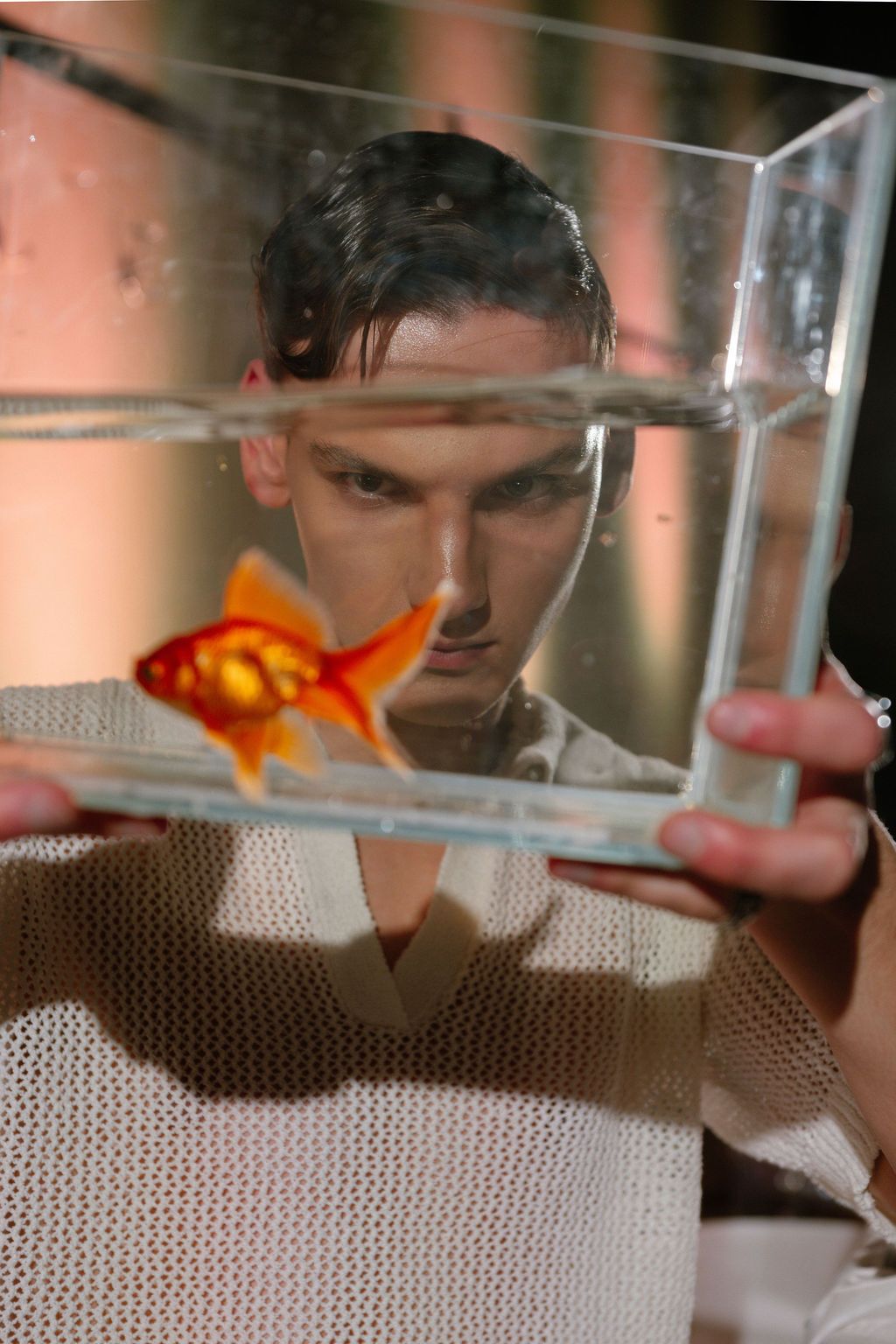 A young man intensely stares at a single goldfish swimming in a clear glass aquarium he holds up.