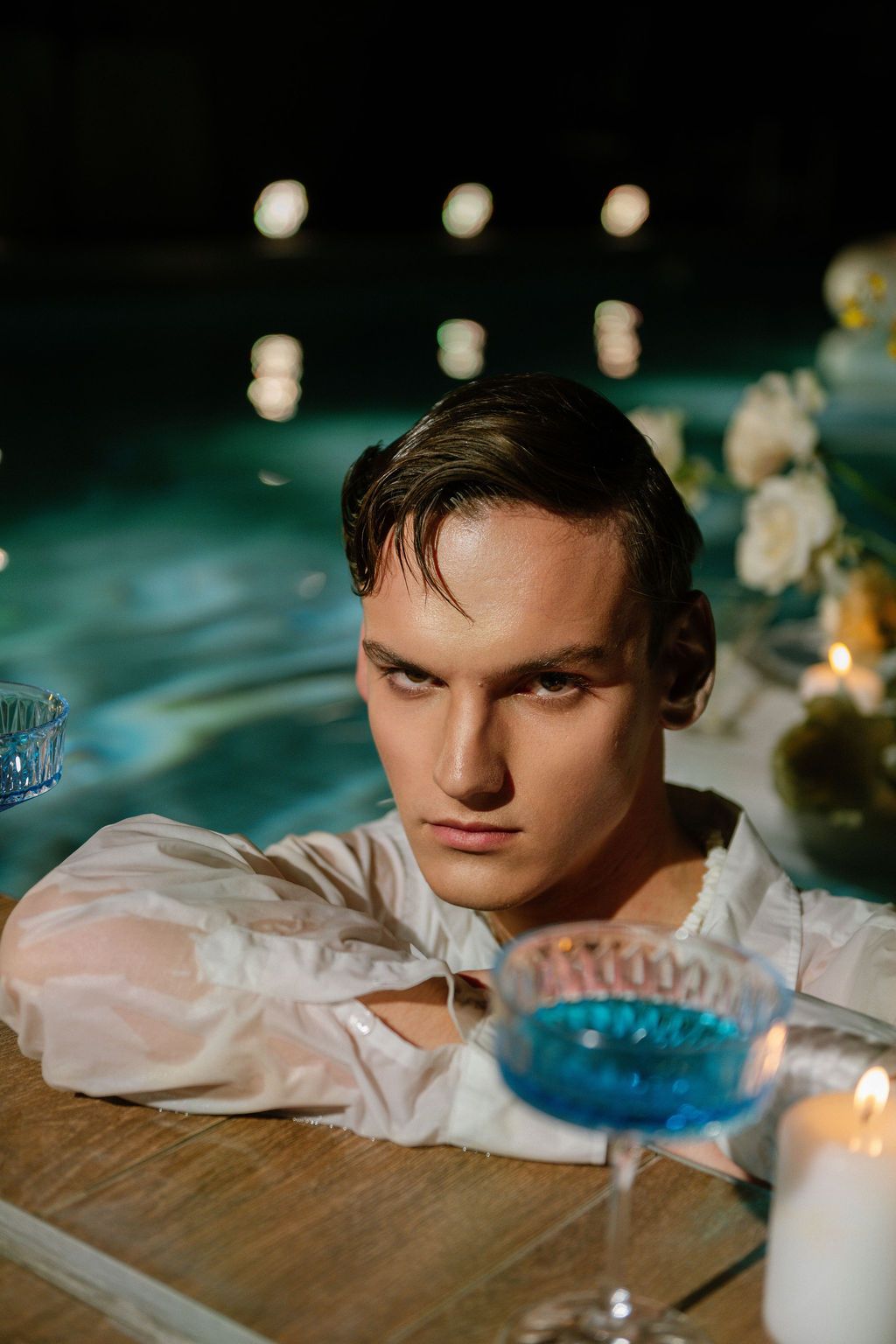 Young man in wet white shirt leaning on pool edge at night with candles and blue cocktail.