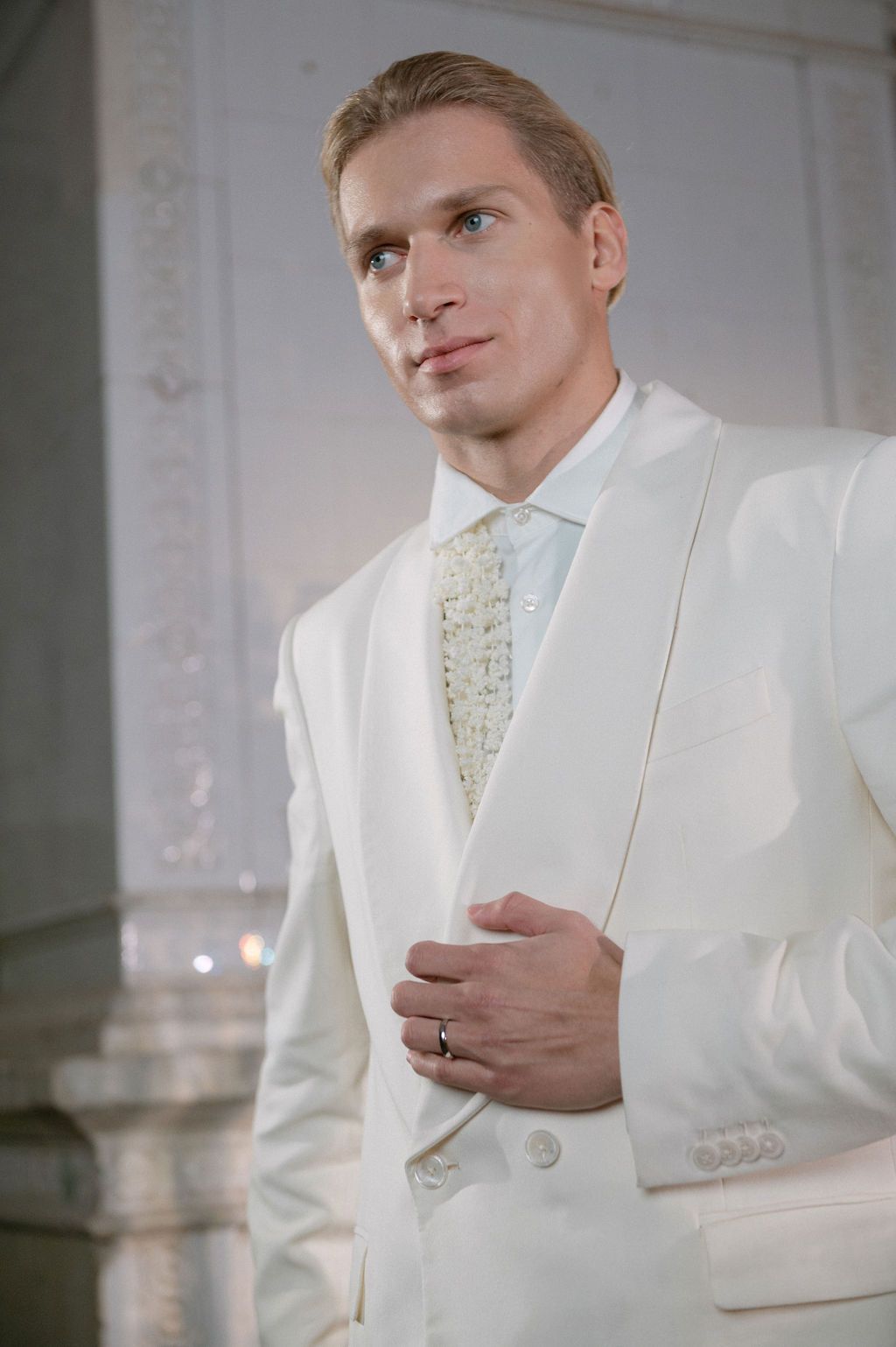 Elegant man in white tuxedo suit with floral tie, posing confidently in a bright, classic interior setting.