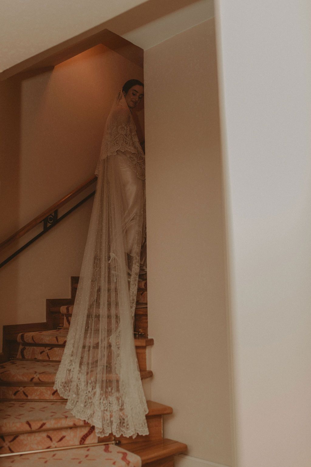 Bride in elegant lace wedding gown and cathedral veil standing on ornate staircase in warm-lit interior.