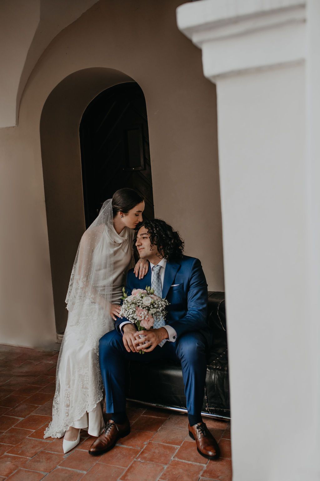 Bride in lace gown leans toward groom in blue suit holding floral bouquet, seated in elegant archway venue.