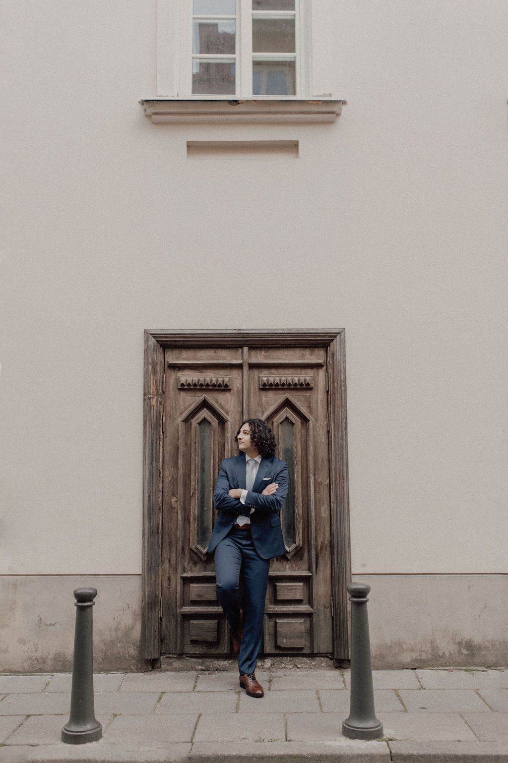 Person in navy suit standing confidently with arms crossed before ornate wooden doors on a city street.