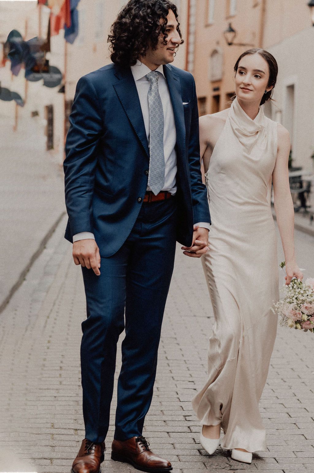 Elegant bride in ivory satin gown holds groom's hand while walking cobblestone street, groom in navy suit and tie.