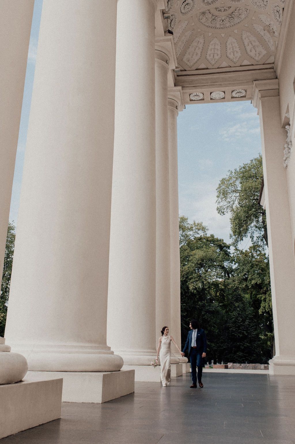 Couple in wedding attire walking between grand white columns of a neoclassical building with lush trees in background.