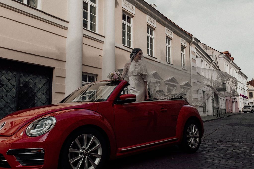 Bride in white gown and veil posing beside a red VW Beetle convertible on a European cobblestone street.