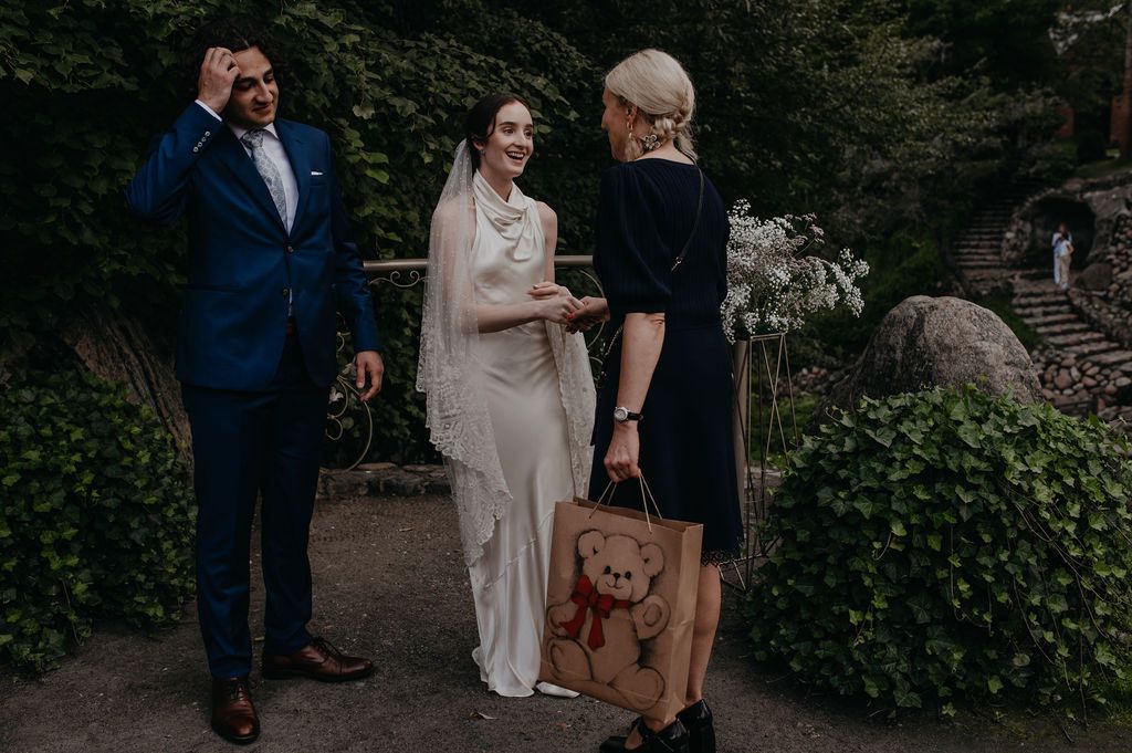 Bride and groom talking with a guest holding a teddy bear gift bag at an outdoor woodland wedding ceremony.