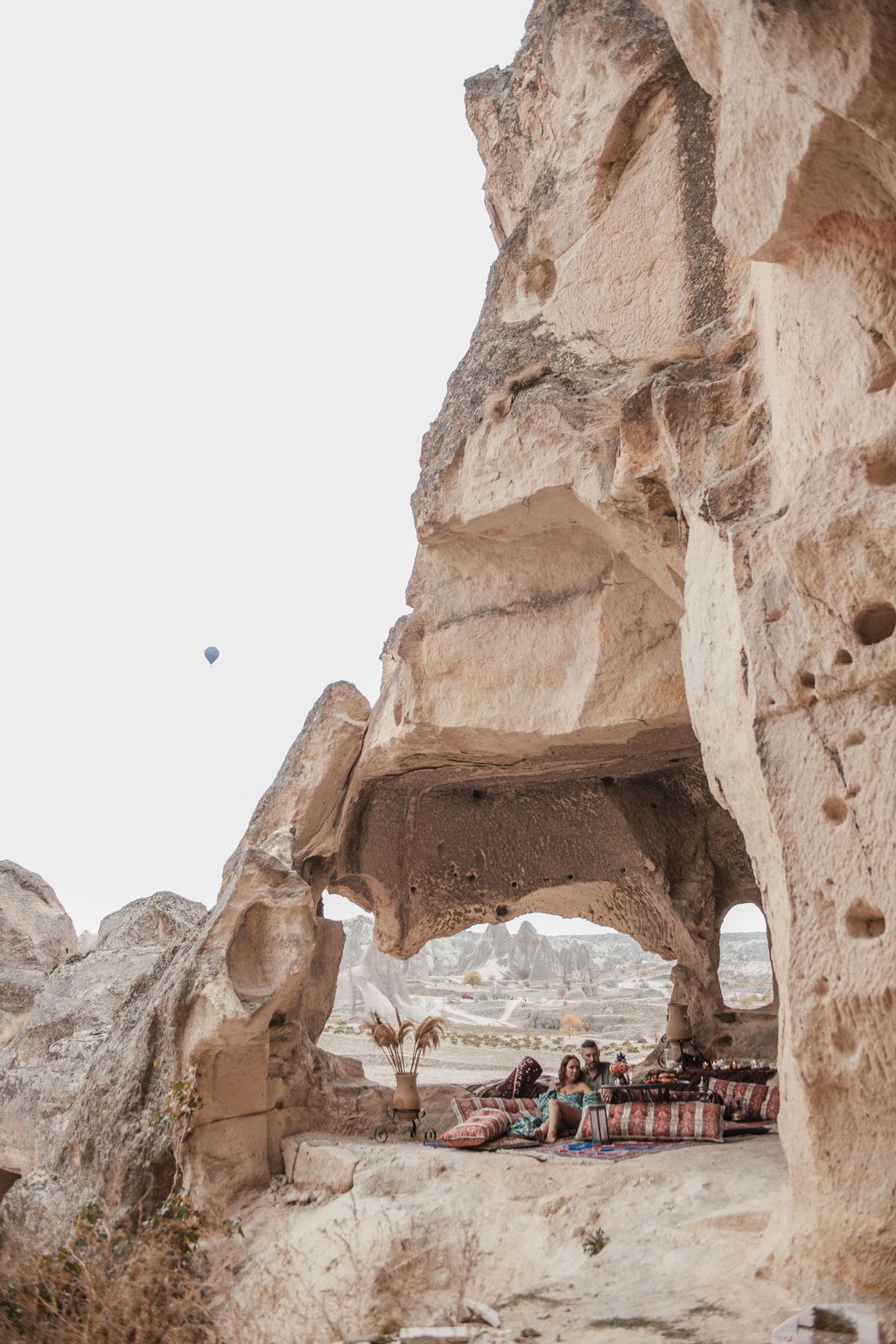 People relaxing in a cave dwelling in Cappadocia, Turkey, with hot air balloons and rocky landscape in background.