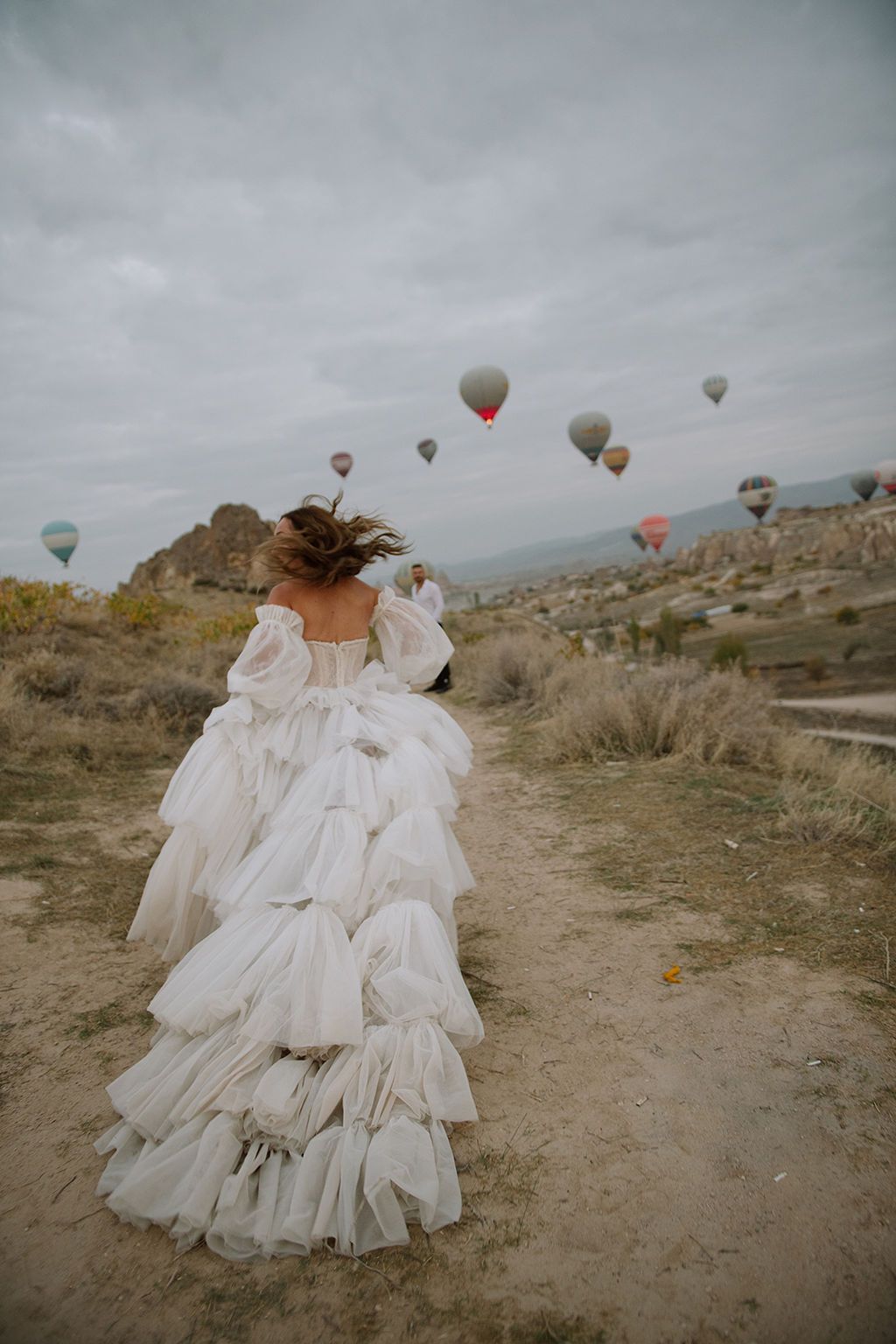 Woman in white ruffled ball gown walks along dirt path with colorful hot air balloons floating in cloudy sky.